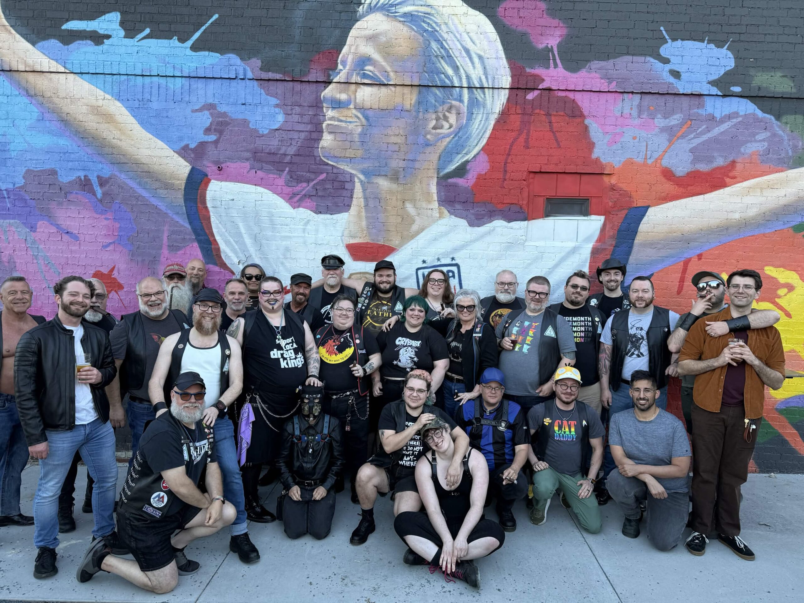 A large group of about 25 people pose together on a sidewalk in front of a colorful mural of a smiling figure with white hair, arms outstretched in a white and red jersey. Most wear black leather vests, harnesses, and pride or leather community pins and patches. People in the front row kneel or sit cross-legged on the ground, while others stand in two rows behind them. Expressions are warm and smiling throughout the group.
